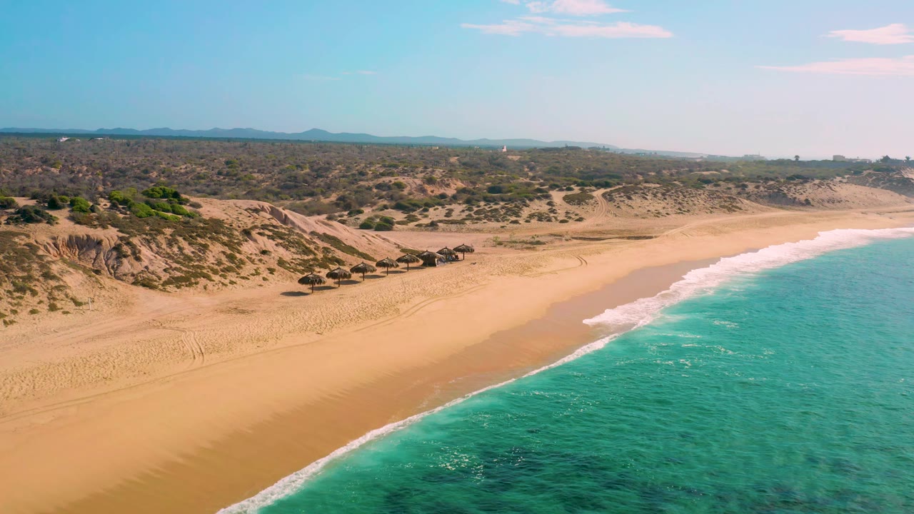 Aerial frame of Shipwrecks Beach showing the shoreline running diagonally, turquoise water on the right, sandy beach and thatched umbrellas near the dunes, with no visible people.