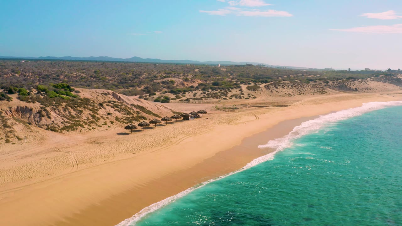Shipwrecks Beach aerial view with pale sand stretching across the frame, teal ocean and breaking surf at right, scrub-covered dunes inland, small thatched umbrellas, and no visible people.
