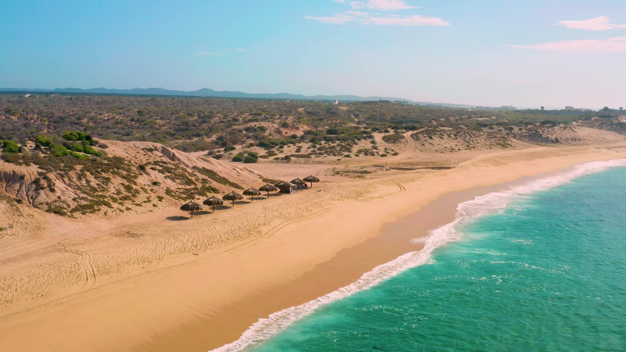 Mid-clip aerial frame over Shipwrecks Beach, with the row of thatched umbrellas lower in the frame, wide sand between dunes and water, gentle surf at right, and no visible people.
