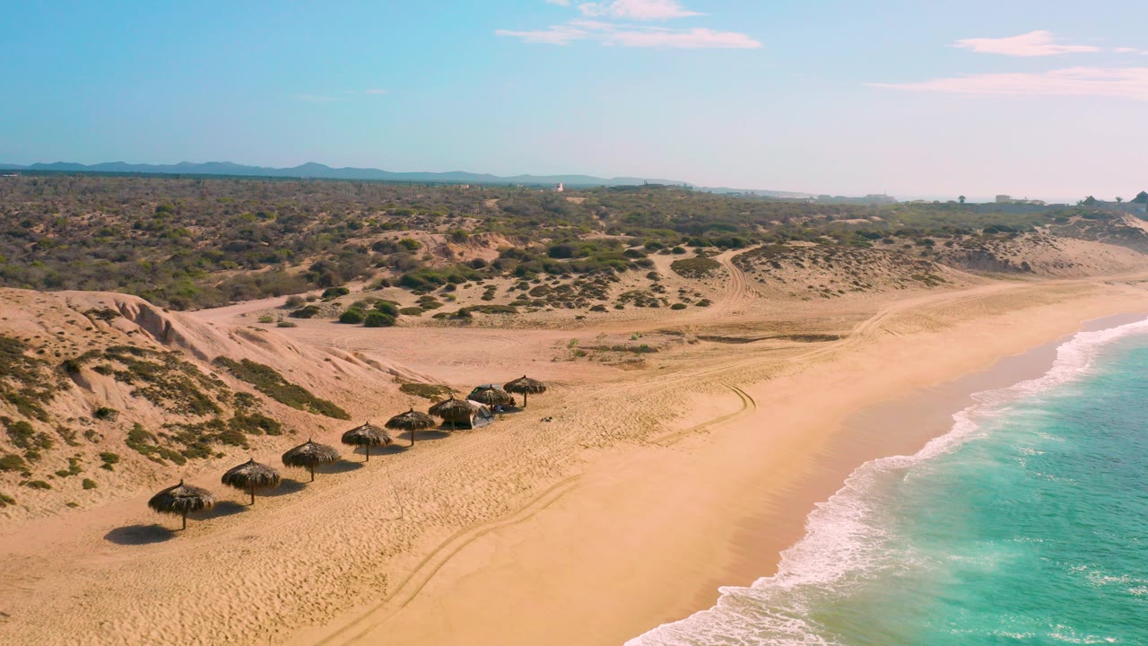 Aerial view of Shipwrecks Beach in Baja California Sur showing palapa shade structures on the sand, scrubby dunes inland, turquoise surf at right, and no visible people.