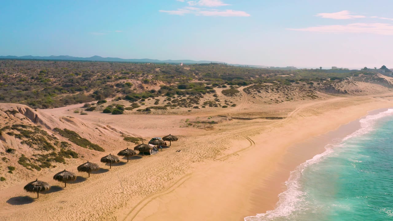 Drone view of Shipwrecks Beach with beach palapas in the lower left, dune ridges and scrub beyond the sand, bright turquoise water at right, and no visible people.