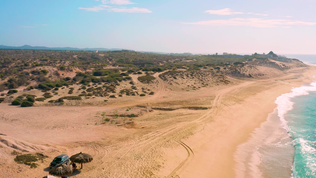 At Shipwrecks Beach in Baja California Sur, an aerial view shows tan sand, white surf along the right edge, scrubby dunes inland, a small shade structure near the lower left, and no visible people.