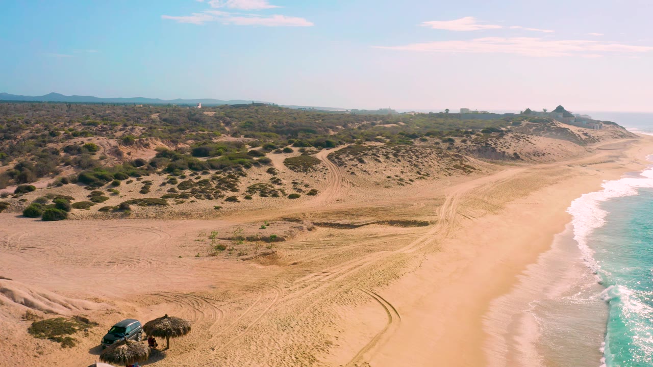 At Shipwrecks Beach in Baja California Sur, the aerial frame keeps the shoreline on the right with white waves, curving tire tracks across the sand, low green brush on dunes, and no visible people.