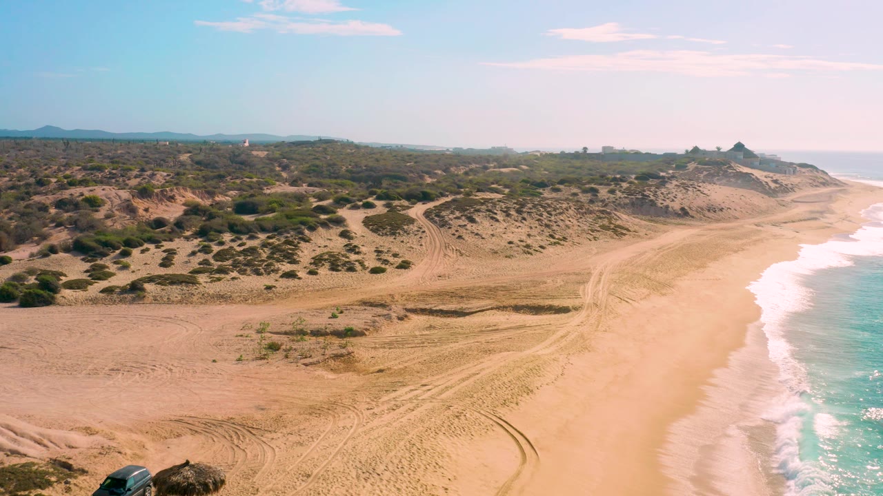 At Shipwrecks Beach in Baja California Sur, surf pushes higher along the right side while broad sandy beach, scrub-covered dunes, faint tracks, and a distant headland remain visible with no visible people.