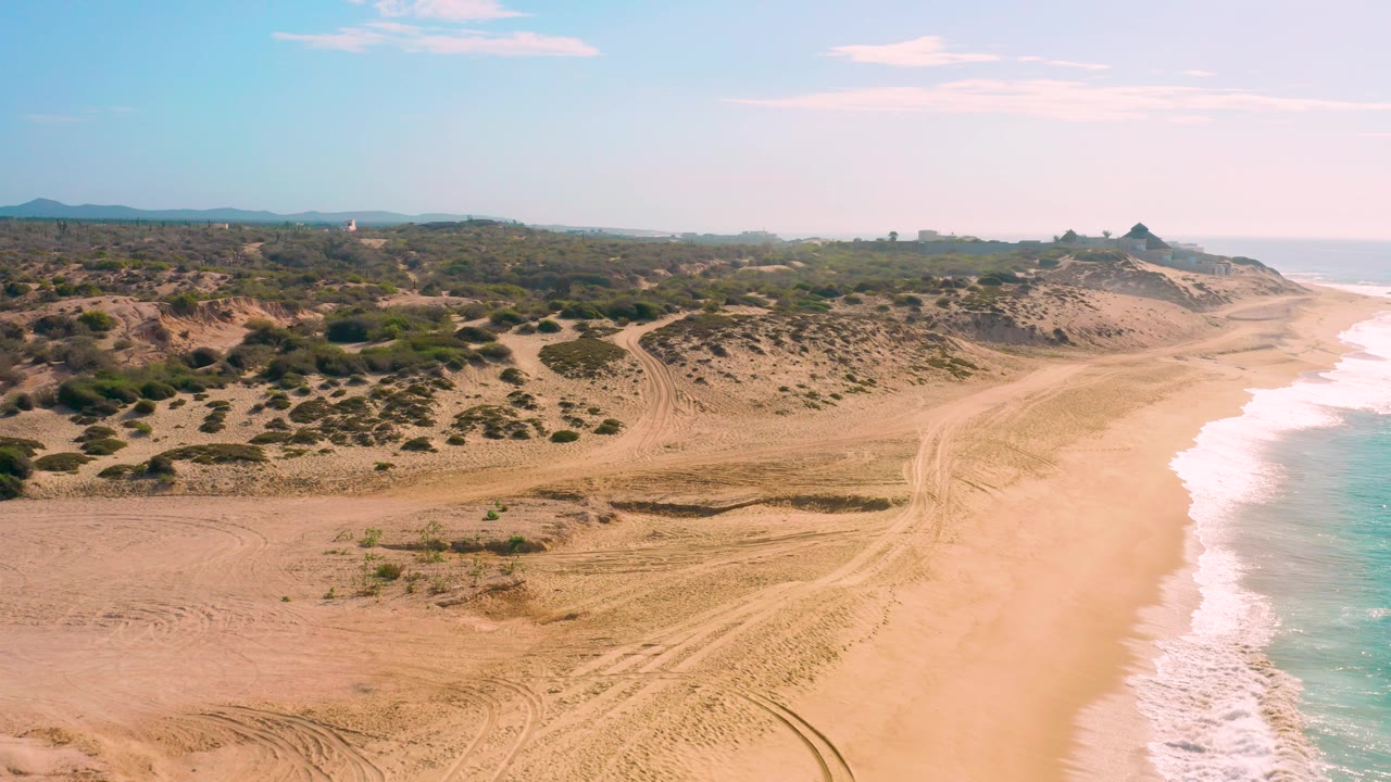 At Shipwrecks Beach in Baja California Sur, the frame shows foamy water along the right, empty tan sand in the foreground, brush patches across the dunes, distant hills, and no visible people.
