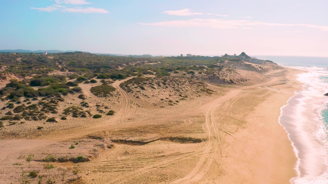 Aerial frame over Shipwrecks Beach in Baja California Sur showing sandy tracks, scrubby dunes, white surf on the right, pale clouds above, and no visible people.