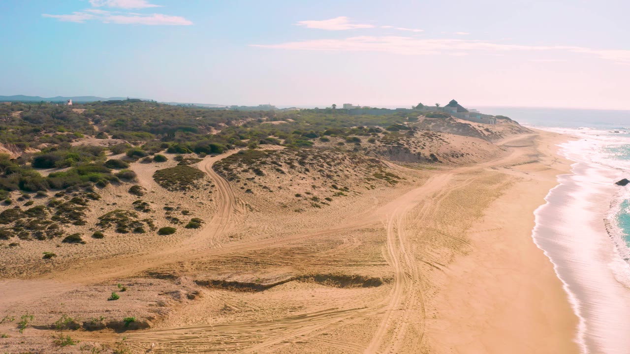 Shipwrecks Beach appears from above with dunes and brush filling the left side, bright foamy waves washing up the right edge, and no visible people on the sand.