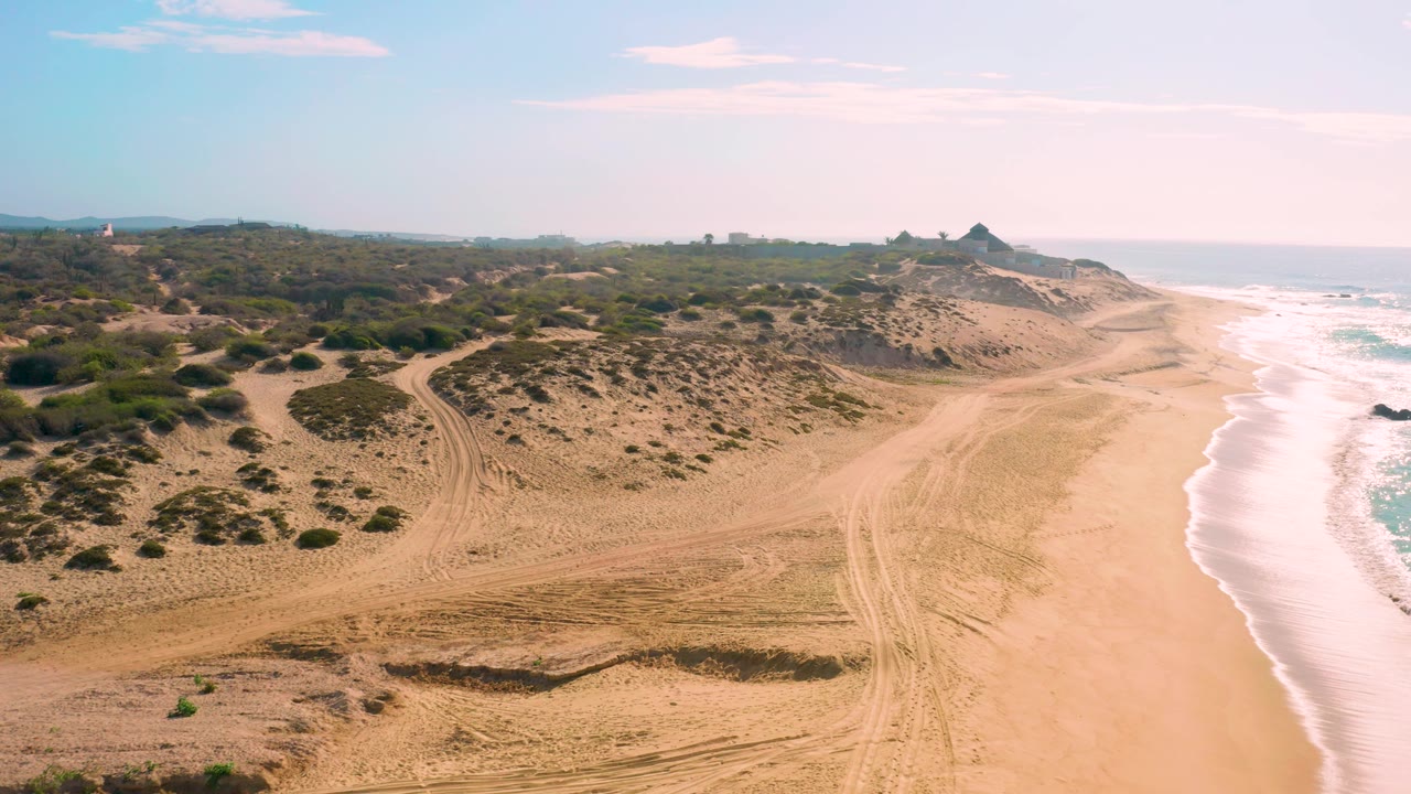 A sunlit aerial view of Shipwrecks Beach shows a broad sandy strip, white surf curling beside it, low scrub on the dunes, distant coastal landforms, and no visible people.