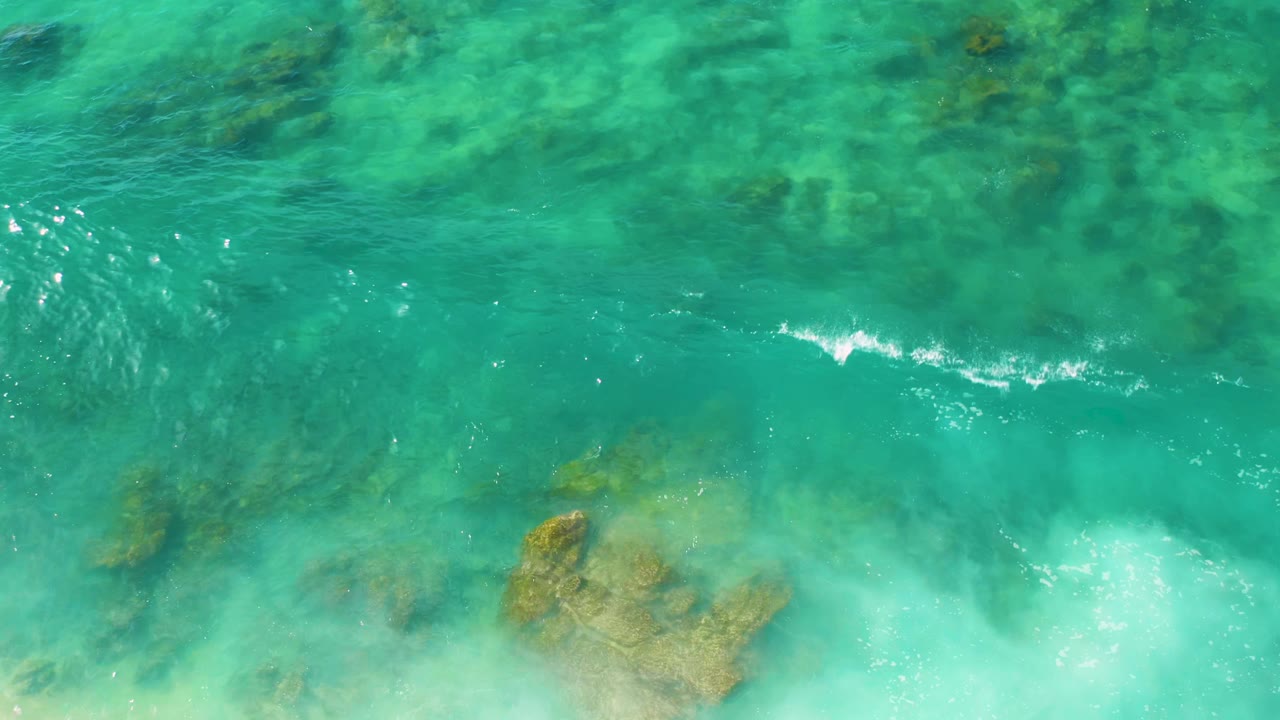 Overhead frame of Shipwrecks Beach water with teal and aquamarine shallows, darker submerged rocks, and soft surf entering near the lower edge, no visible people.