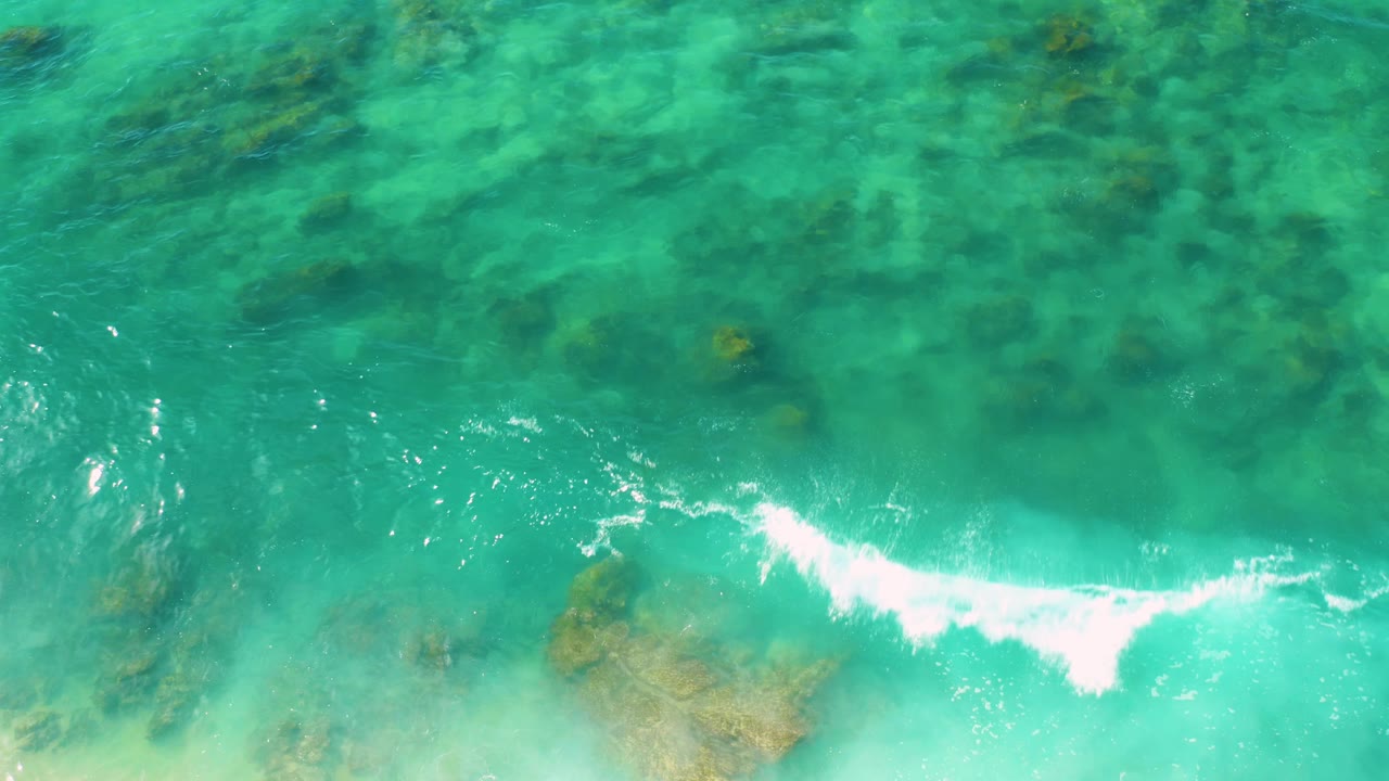Aerial view at Shipwrecks Beach with bright turquoise water, a curved white foam break, and clustered submerged rocks beneath the surface, no visible people.