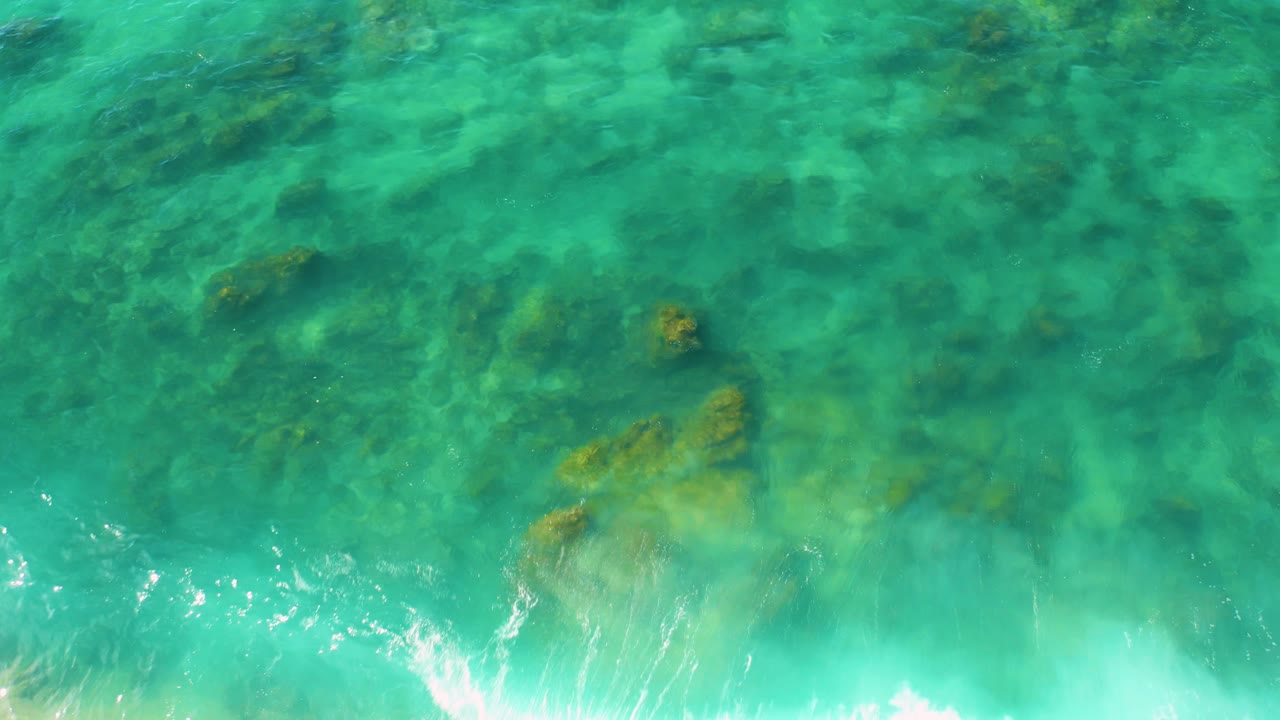 Top-down Shipwrecks Beach frame showing green-blue water, multiple submerged rock patches, pale seabed areas, and a small band of white surf at the lower left, no visible people.