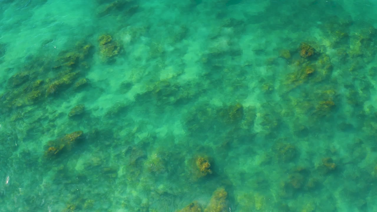 Aerial frame over shallow blue-green ocean at Shipwrecks Beach, with darker reef patches visible under the surface and no visible people.