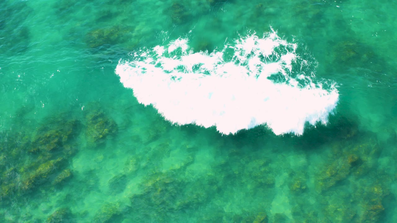 Overhead view of turquoise water at Shipwrecks Beach in Baja California Sur, with a white foam band crossing the upper frame, dark algae patches below the surface, and no visible people.