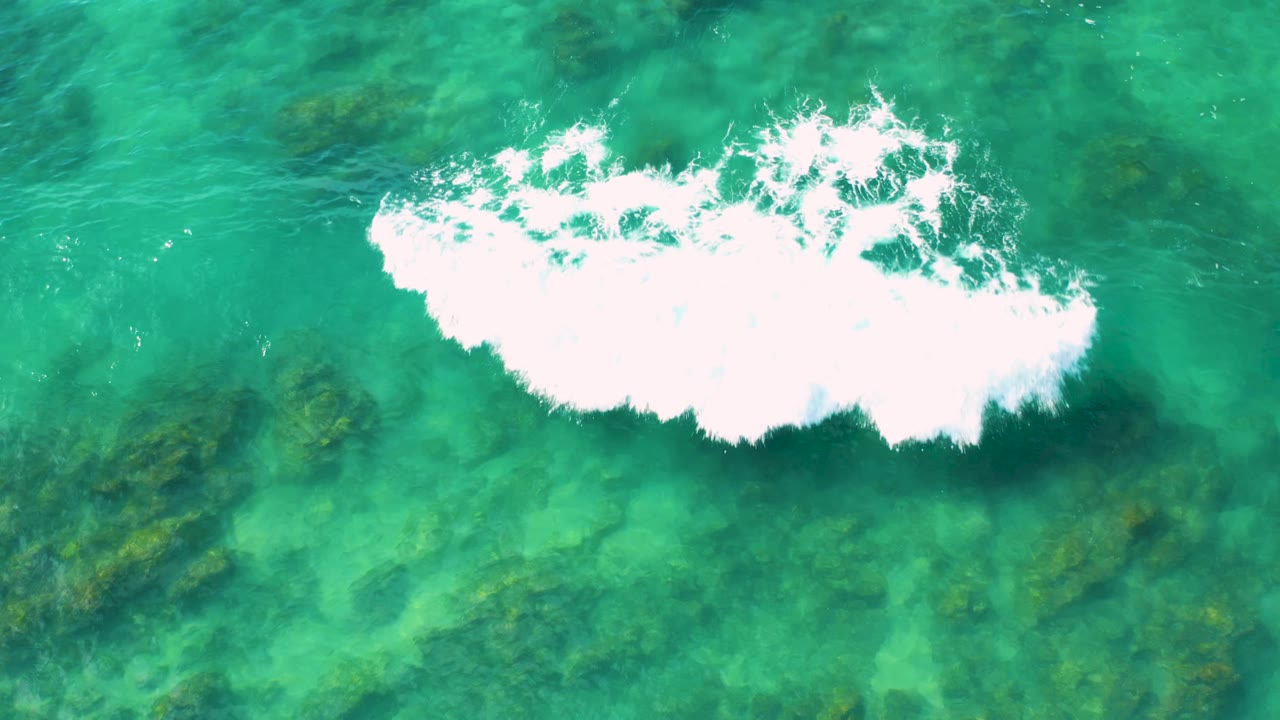 Aerial frame at Shipwrecks Beach showing green-blue clear ocean, a broad white foam patch near the top edge, scattered darker algae areas, and no visible people.