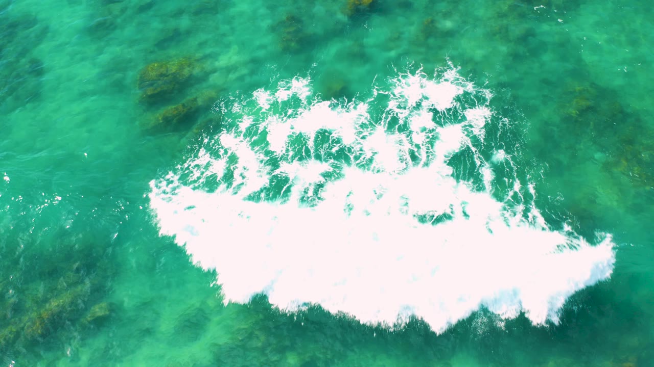 Top-down Shipwrecks Beach view with bright white surf foam spreading across the right side of turquoise water, algae patches visible beneath, and no visible people.