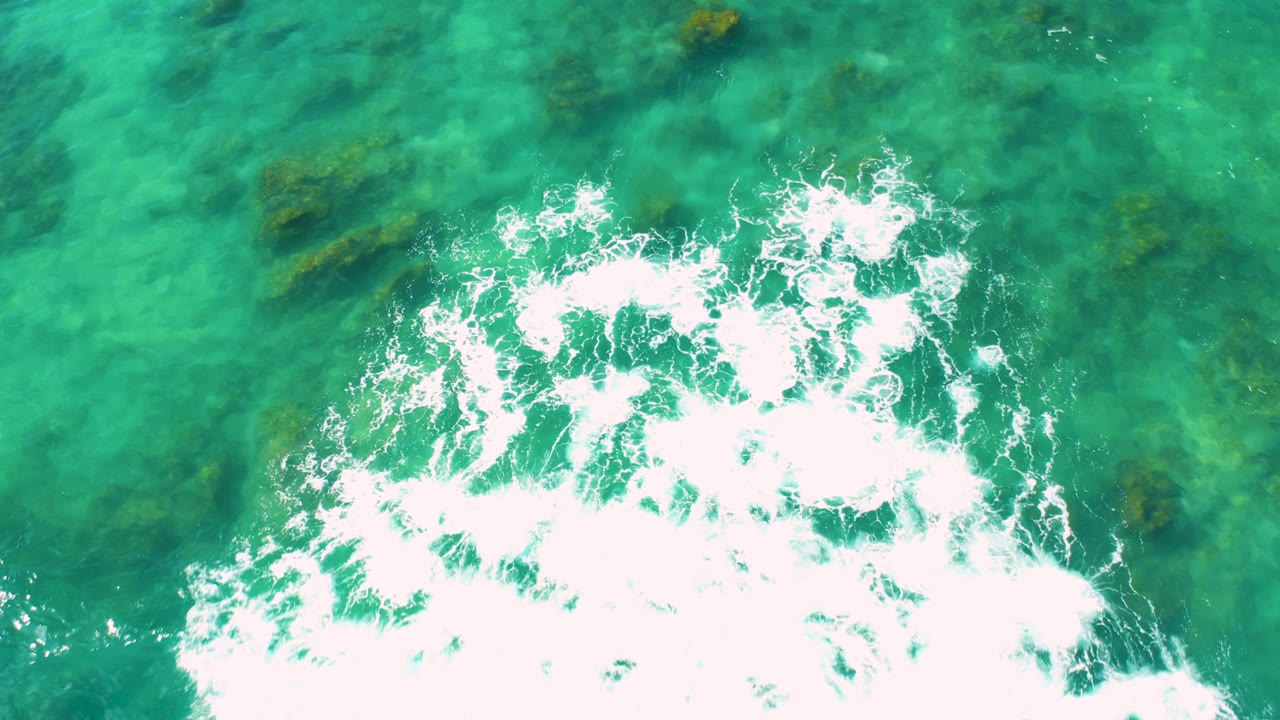 Overhead Baja California Sur ocean frame with a foamy wave edge entering from the lower left, clear green water across the frame, algae patches, and no visible people.