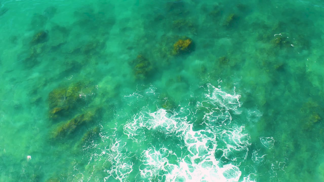 Aerial view of clear blue-green water at Shipwrecks Beach, Baja California Sur, with a bright foam patch near the lower center and no visible people.