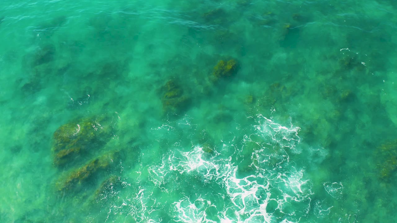 Overhead frame of Shipwrecks Beach water with scattered submerged rock patches, white foam along the lower edge, and no visible people.