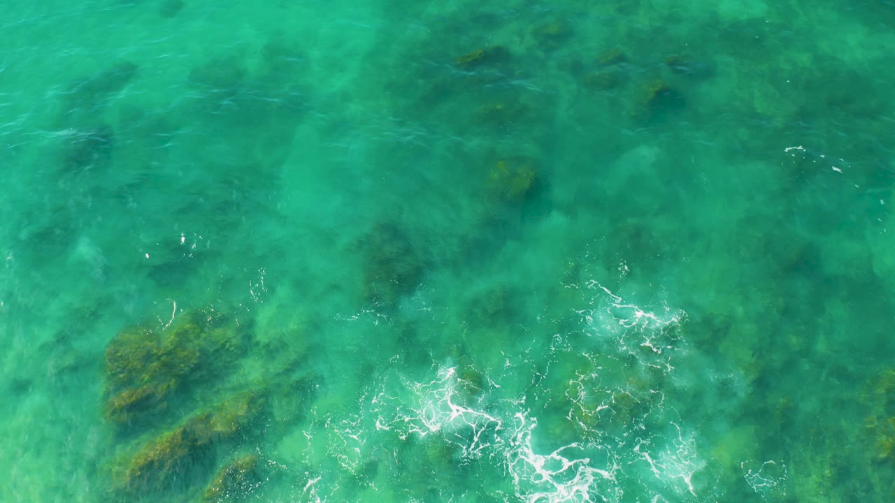 Top-down view at Shipwrecks Beach showing calmer turquoise water, muted reef shadows, a small white foam streak, and no visible people.