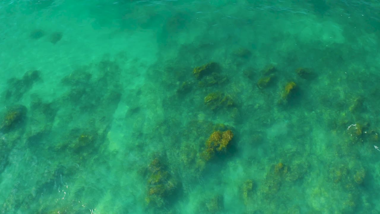 Clear green-blue water at Shipwrecks Beach in Baja California Sur reveals a denser cluster of submerged rocks and coral near the center, with no visible people.