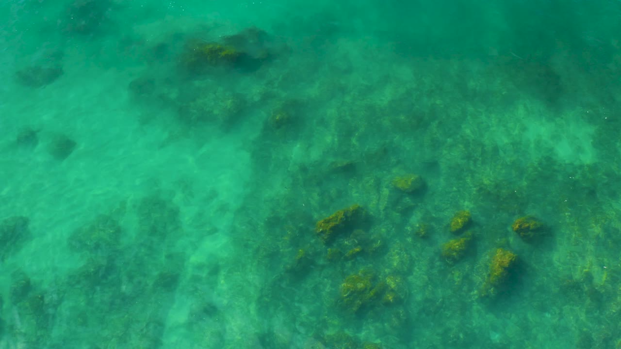 A patch of dark and yellow-green rocks sits below bright turquoise water at Shipwrecks Beach in Baja California Sur, with no visible people.