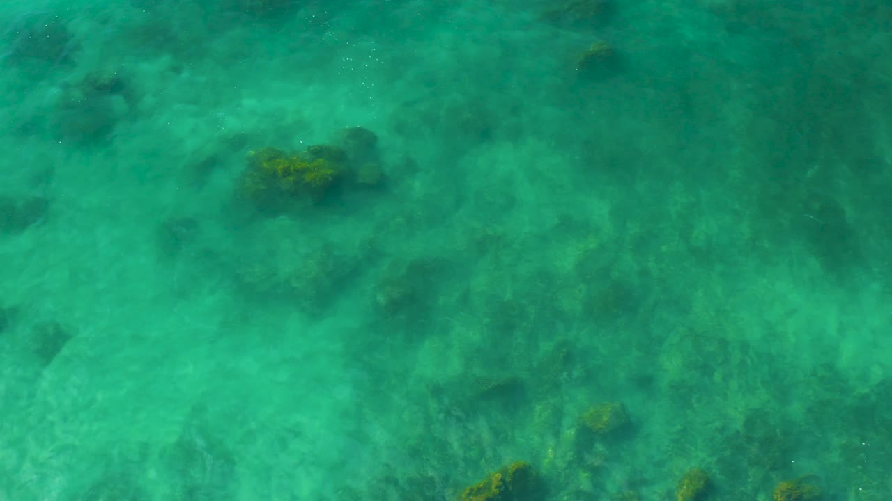 A top-down water plate shows scattered rock shapes under translucent turquoise water at Shipwrecks Beach, Baja California Sur, with no visible people.