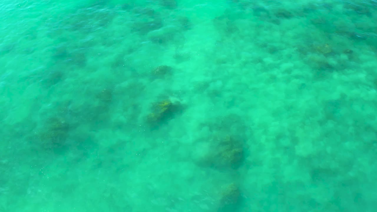 Green-blue sea surface viewed from above at Shipwrecks Beach, with darker underwater rock or reef shapes softened by ripples and no visible people.