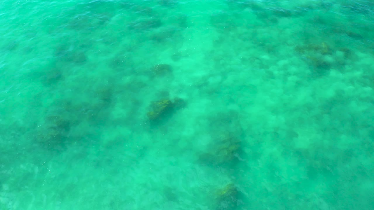 Aerial-looking view over shallow turquoise water in Baja California Sur, with scattered darker seabed patches and no people, boats, or shoreline visible.