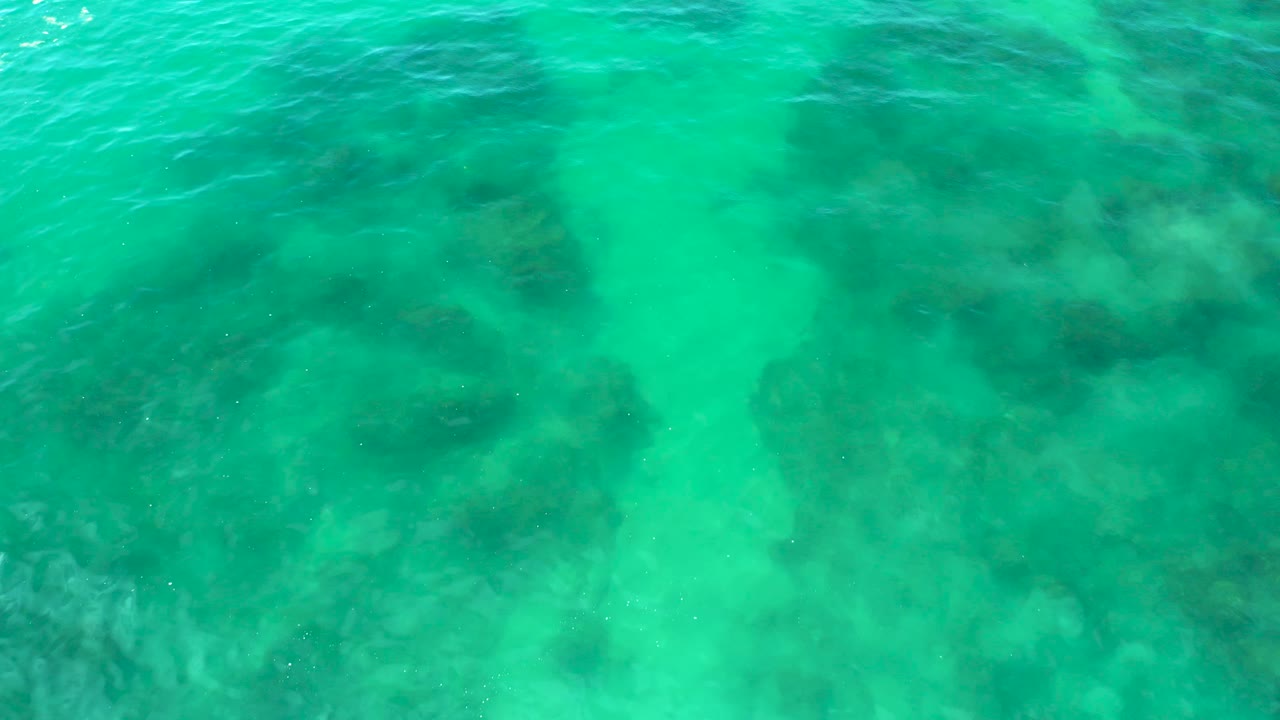Straight-down aerial view of clear water at Shipwrecks Beach, Baja California Sur, showing pale lagoon floor between dark submerged rock patches and no visible people.