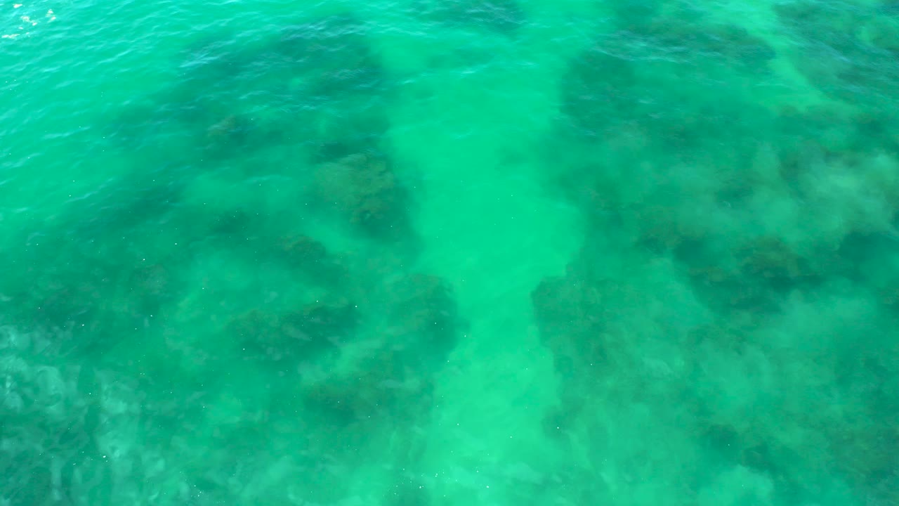 Turquoise shallow water covers broad dark rock areas beneath the surface at Shipwrecks Beach, with small ripples near the top of frame and no visible people.