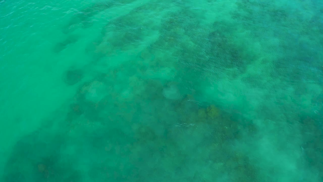 Clear green ocean water at Shipwrecks Beach, Baja California Sur, with a broad darker underwater formation centered beneath faint surface ripples and no visible people.