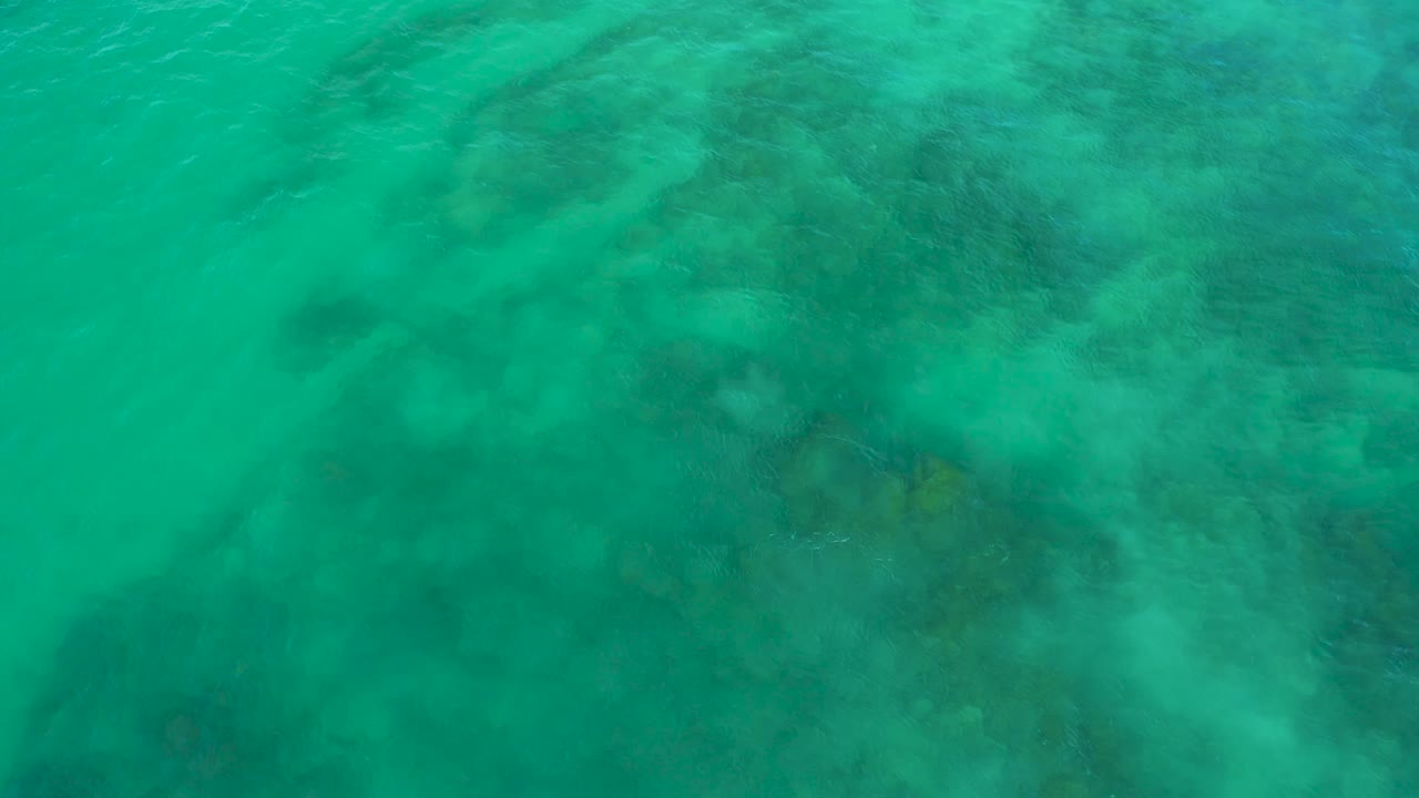 Aerial view of turquoise-green ocean at Shipwrecks Beach, Baja California Sur, where darker seabed areas sit beneath a lightly rippled surface with no visible people.
