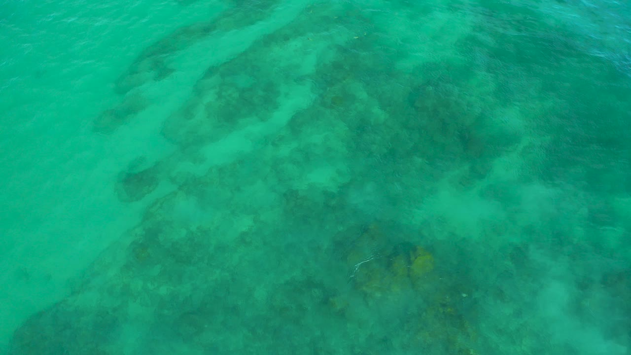 Overhead view of turquoise water at Shipwrecks Beach in Baja California Sur, with dark submerged reef shapes under the surface and no visible people.