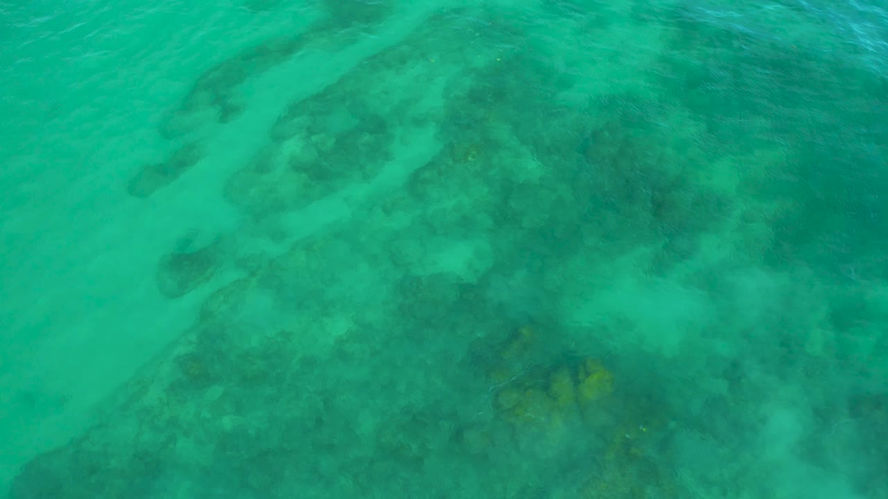A top-down frame from Shipwrecks Beach shows clear shallow water, uneven dark reef areas, and pale sand showing through, with no visible people.