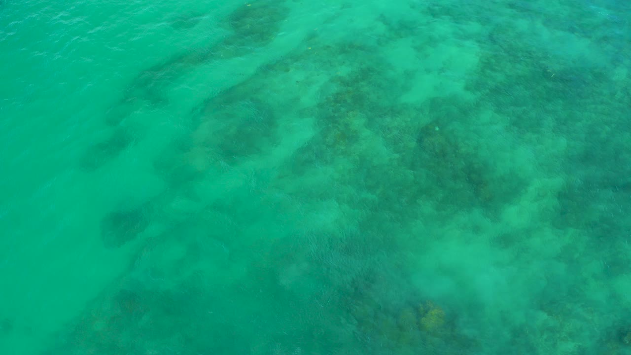 Top-down view of turquoise water at Shipwrecks Beach in Baja California Sur, with darker submerged rock or reef shapes under the surface and no visible people.