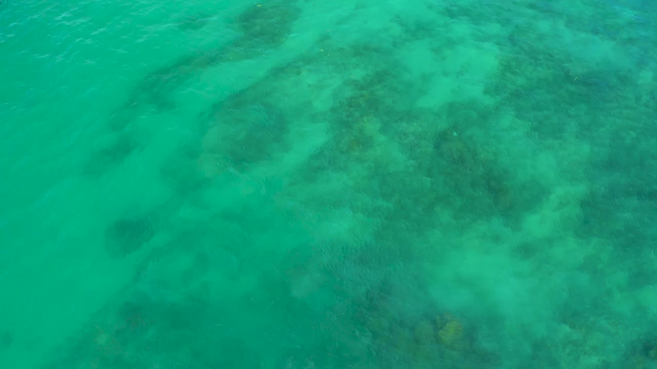 Aerial-looking top-down water frame from Shipwrecks Beach, with mottled darker seabed shapes softened by turquoise surface ripples and no visible people.
