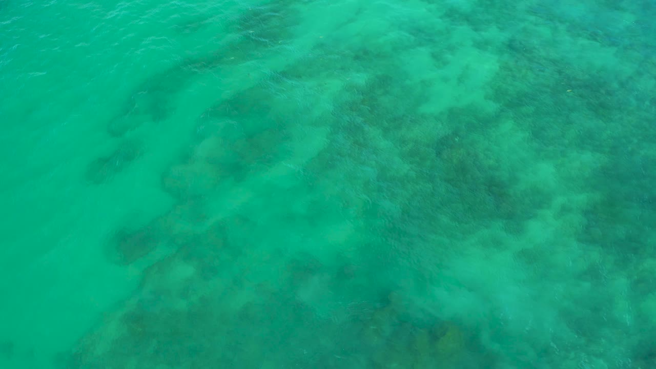 Top-down frame over Shipwrecks Beach water, where broad darker underwater patches sit beneath a bright turquoise surface with no visible people or objects.