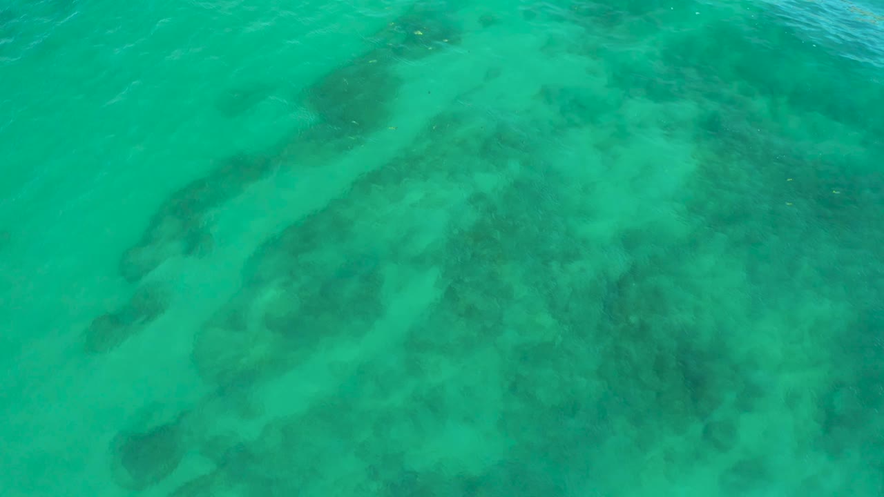 Clear green-blue water at Shipwrecks Beach shows mottled coral or rock patches beneath light surface ripples, with no visible people or objects.