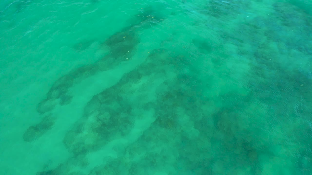 Shallow turquoise water at Shipwrecks Beach is crossed by soft wavelets over dark seabed areas, with no shoreline, boats, or people visible.