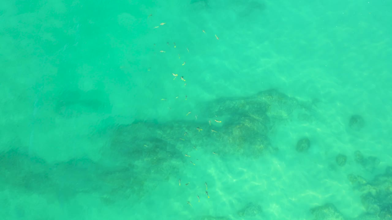 Top-down turquoise water at Shipwrecks Beach, Baja California Sur, with faint dark underwater patches and a small round darker spot to the right, with no visible people.