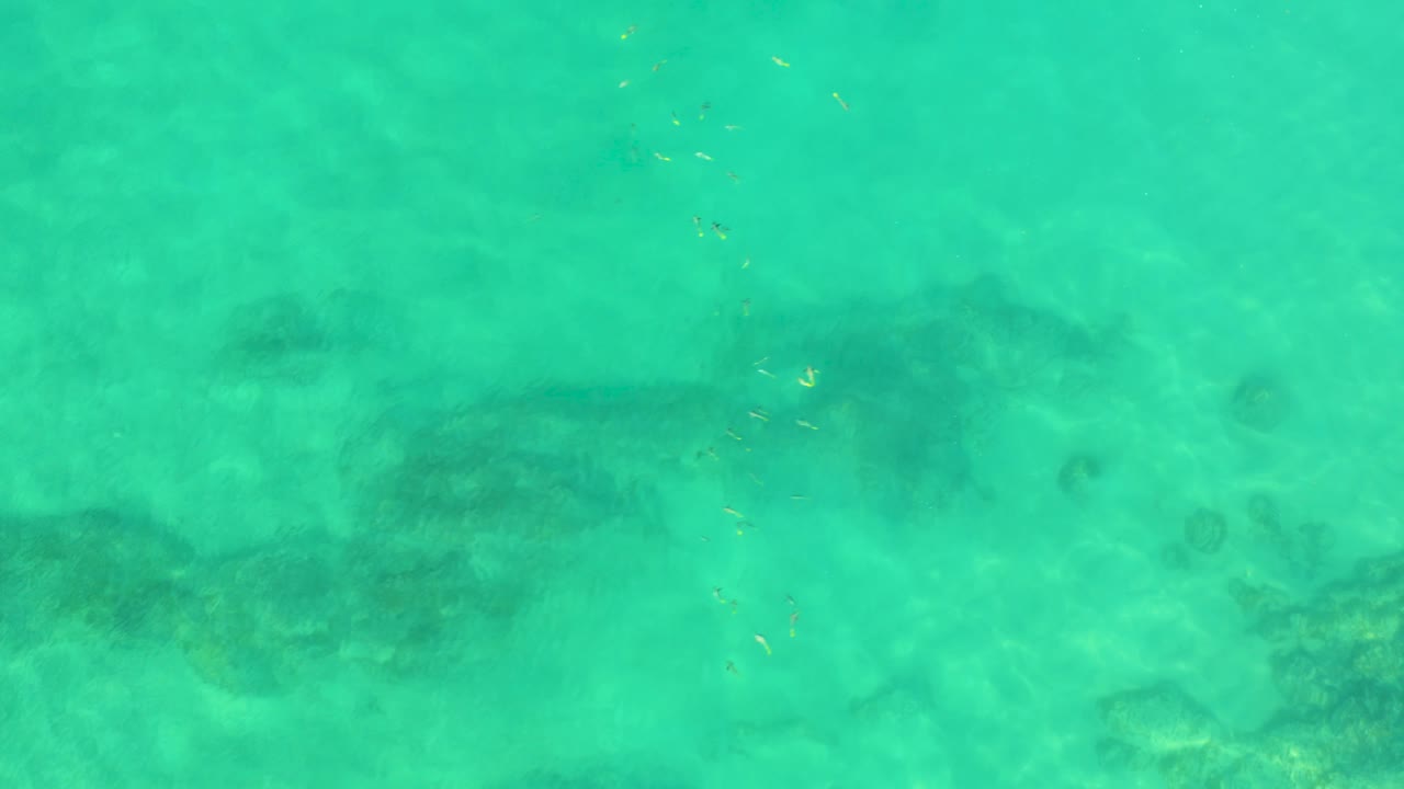 High aerial view of pale turquoise water at Shipwrecks Beach, Baja California Sur, with a dark underwater ridge crossing the lower left and no visible people.