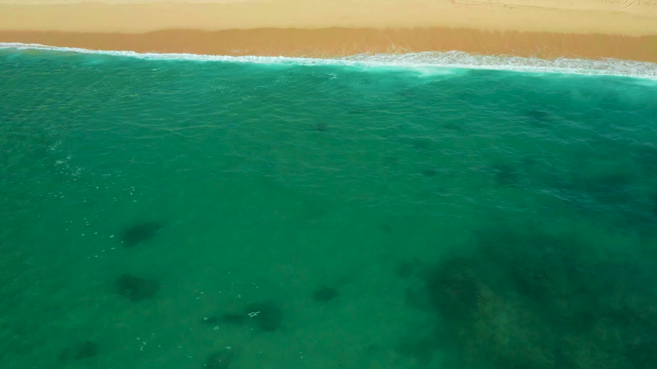 Aerial view over Shipwrecks Beach in Baja California Sur, with turquoise water, a narrow line of white surf, pale sand at the top, dark submerged patches, and no visible people.