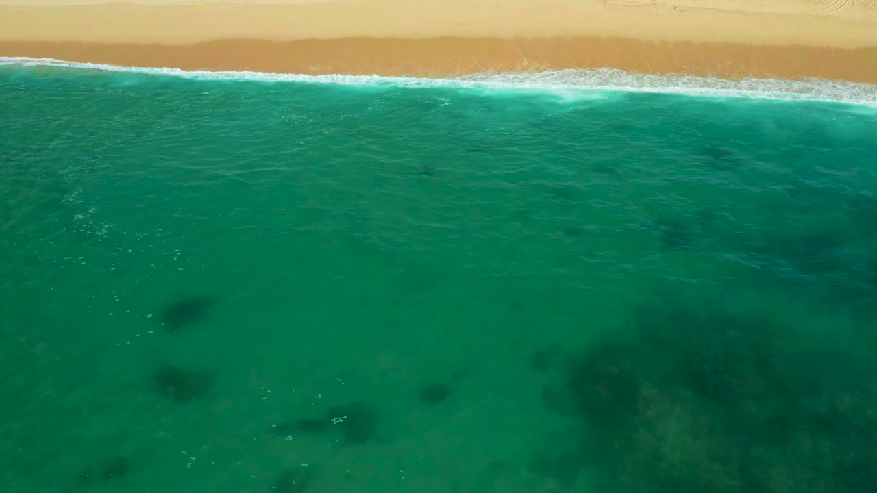 Aerial frame at Shipwrecks Beach showing green-blue water filling most of the view, soft waves breaking along the upper sandbar, scattered dark underwater shapes, and no visible people.