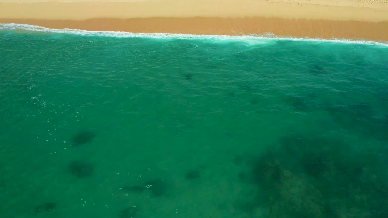 Overhead coastal view at Shipwrecks Beach, with a continuous foam line against pale sand, clear teal water below, darker reef or rock patches under the surface, and no visible people.