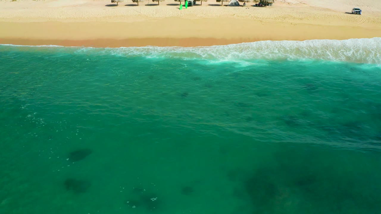 Aerial frame at Shipwrecks Beach, Baja California Sur, with blue-green water dominating the view, a brighter foam band near shore, and beach structures along the upper sand, with no visible people.
