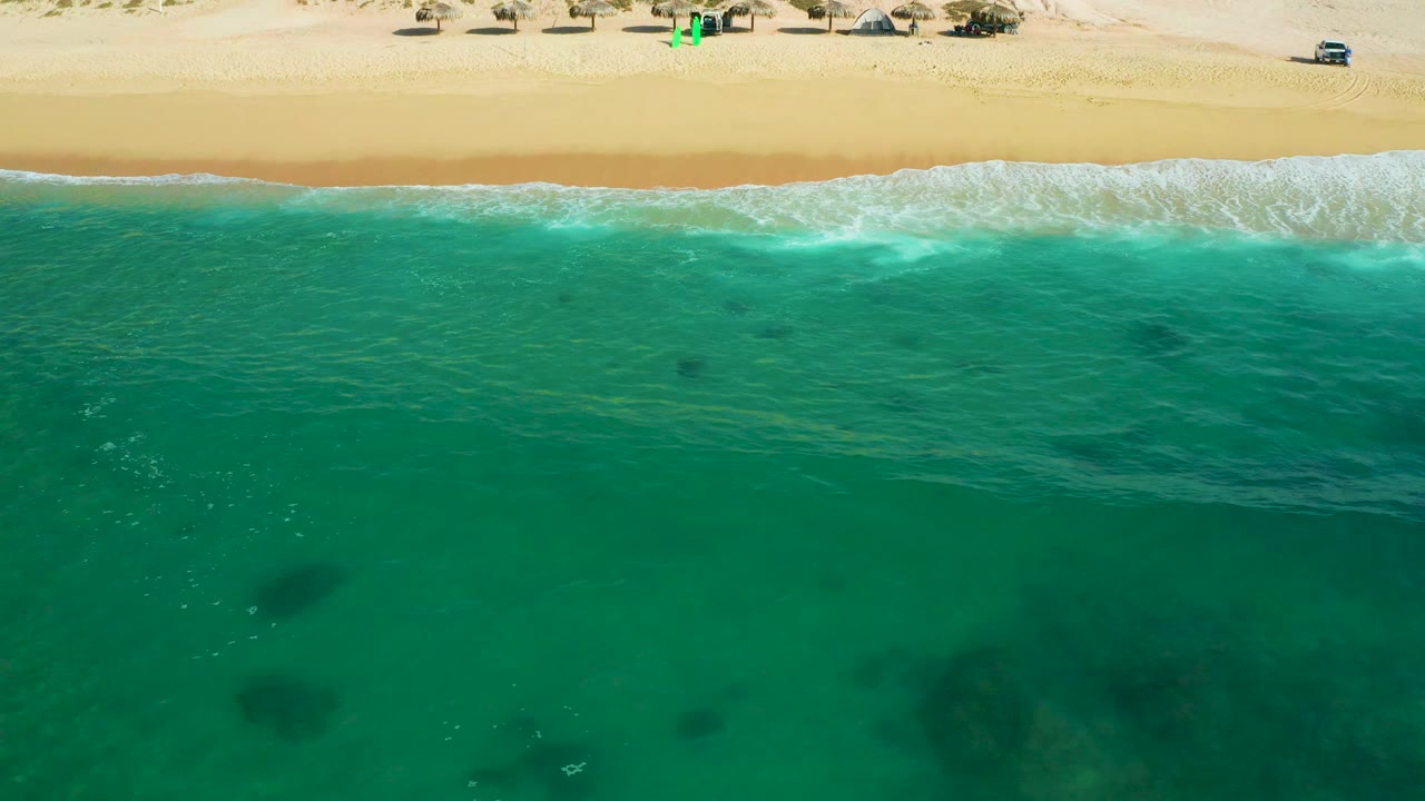 Overhead view at Shipwrecks Beach, Baja California Sur, showing a longer strip of white wash across the shoreline, deep turquoise water in the foreground, and empty sunlit sand above, with no visible people.