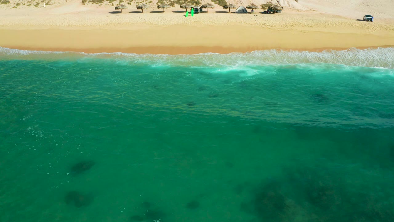 Aerial view of Shipwrecks Beach, Baja California Sur, where a low wave spreads white foam along the sand and green-blue water fills the lower frame, with no visible people.