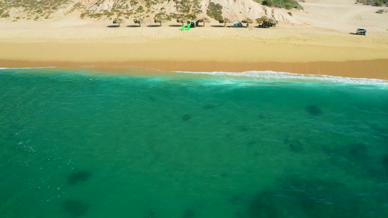 Aerial frame over Shipwrecks Beach in Baja California Sur, with turquoise water in front, a thin white surf line, sandy beach, dunes with scrub, small vehicles and shade structures, and no visible people.
