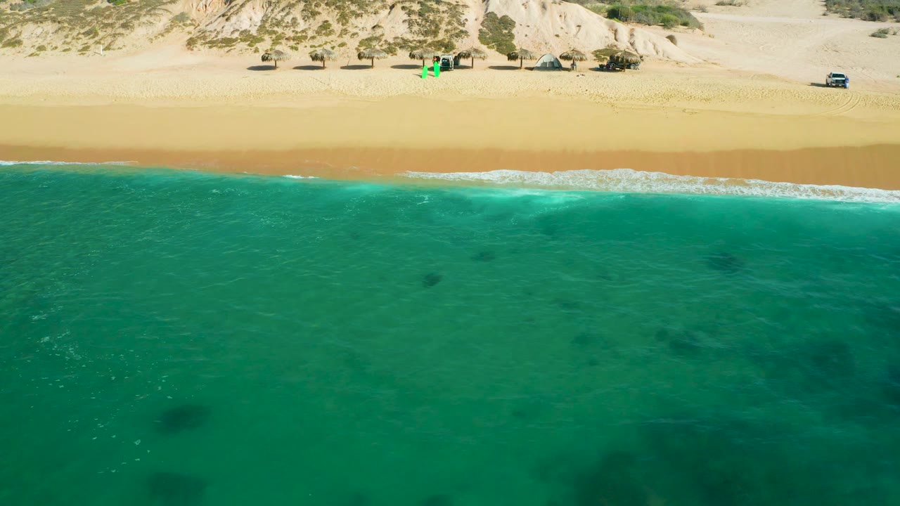 Drone-style frame from Shipwrecks Beach showing broad calm water in the foreground, small darker patches under the surface, a low foamy shore break, open sand, dune vegetation, vehicles, and no visible people.