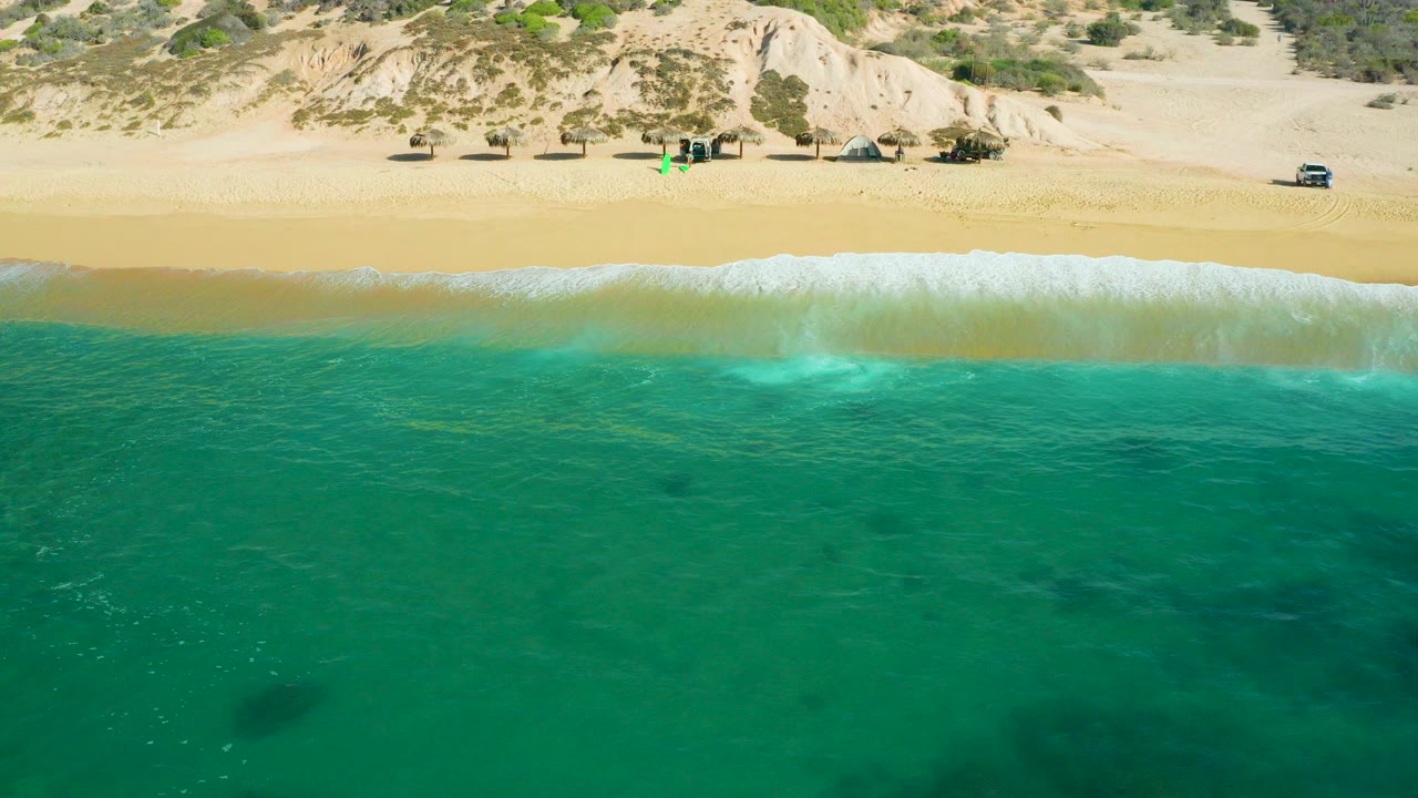 Aerial frame at Shipwrecks Beach, Baja California Sur, showing turquoise water below a white surf line, golden sand, pale dunes, scrub plants, small beach structures, and no visible people.